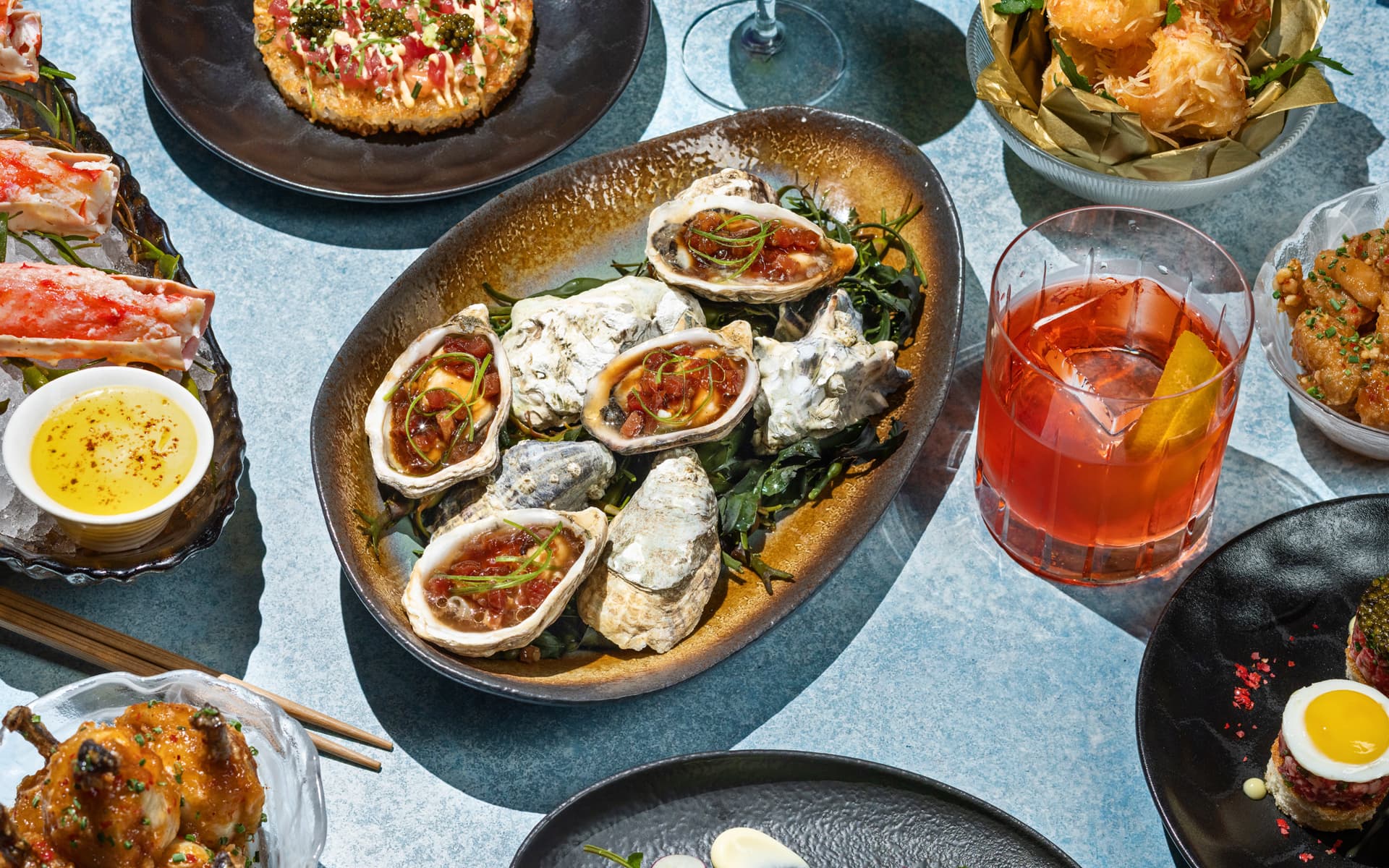 A spread of upscale seafood and appetizers on a blue table, including oysters on the half shell with sauce, crab legs with melted butter, fried shrimp, tartare with quail egg, and a red cocktail with an orange twist.