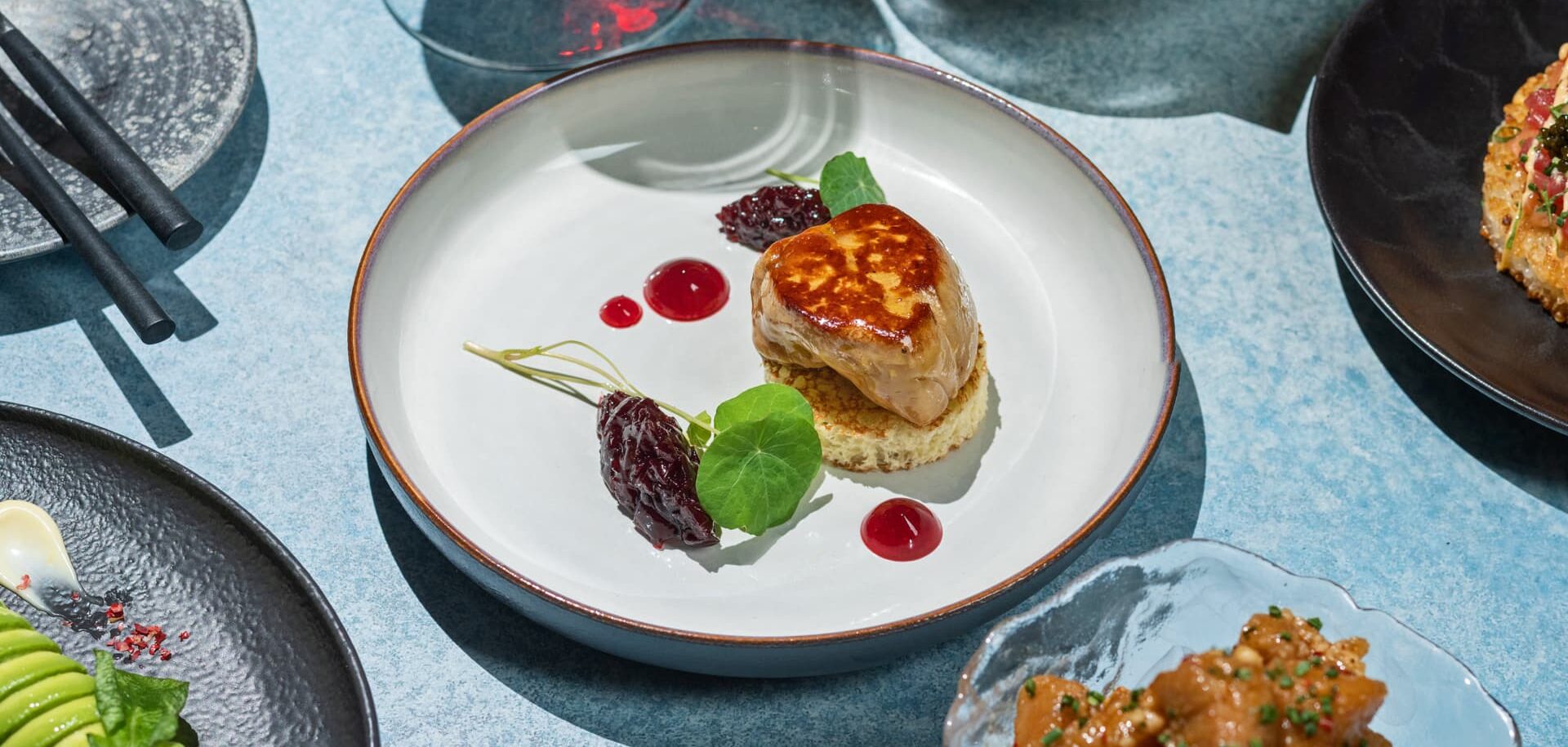 A plated gourmet dish featuring seared foie gras on a round toast, garnished with red fruit compote, microgreens, and small drops of sauce, surrounded by other fine-dining plates on a blue table.