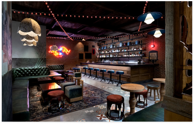 A dimly lit bar with exposed brick walls, leather seating, a stocked liquor shelf, eclectic stools, and a neon tiger sign adding a vibrant touch.