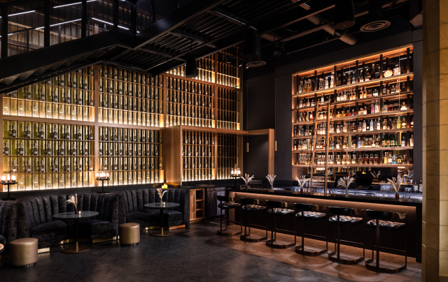 A luxurious bar with floor-to-ceiling illuminated liquor shelves, dark velvet seating, gold accents, and a sleek counter with bar stools.