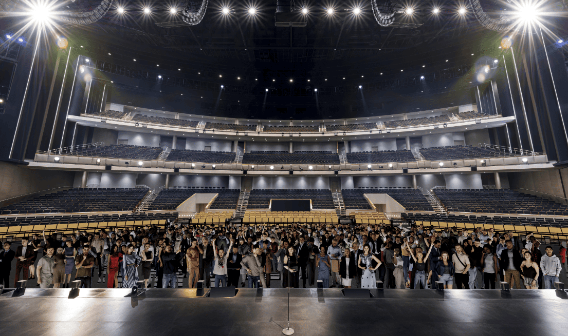 Large theater interior with three levels of seating and a crowd gathered in front of the stage under bright stage lights.

