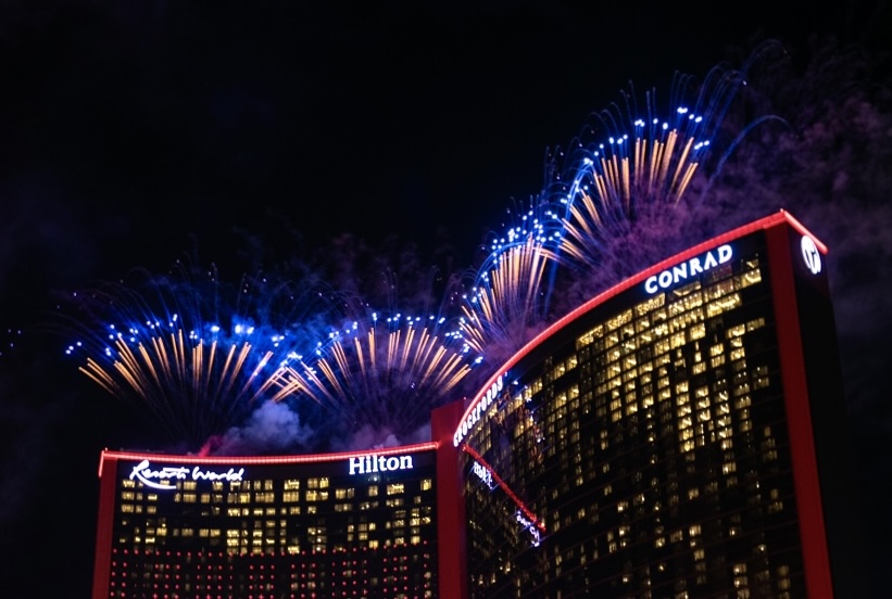 Fireworks display above Resorts World towers at night, featuring Hilton and Conrad buildings outlined in red lighting.