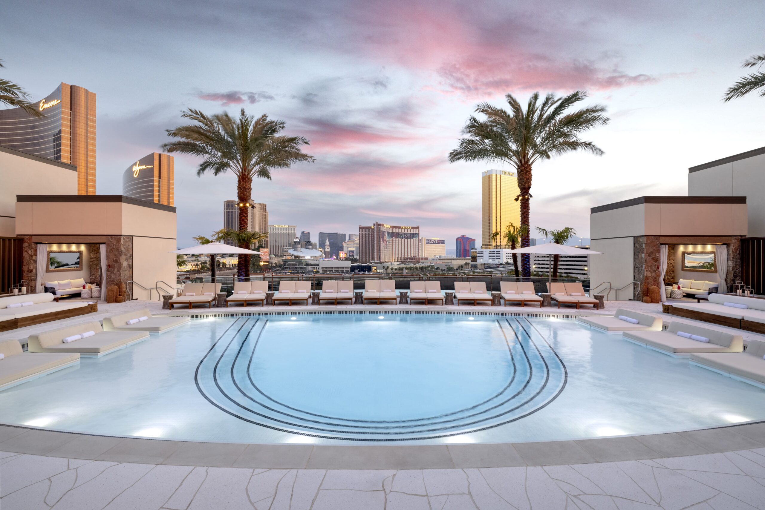 Luxurious rooftop pool with lounge chairs, cabanas, and palm trees overlooking the Las Vegas Strip at sunset. 