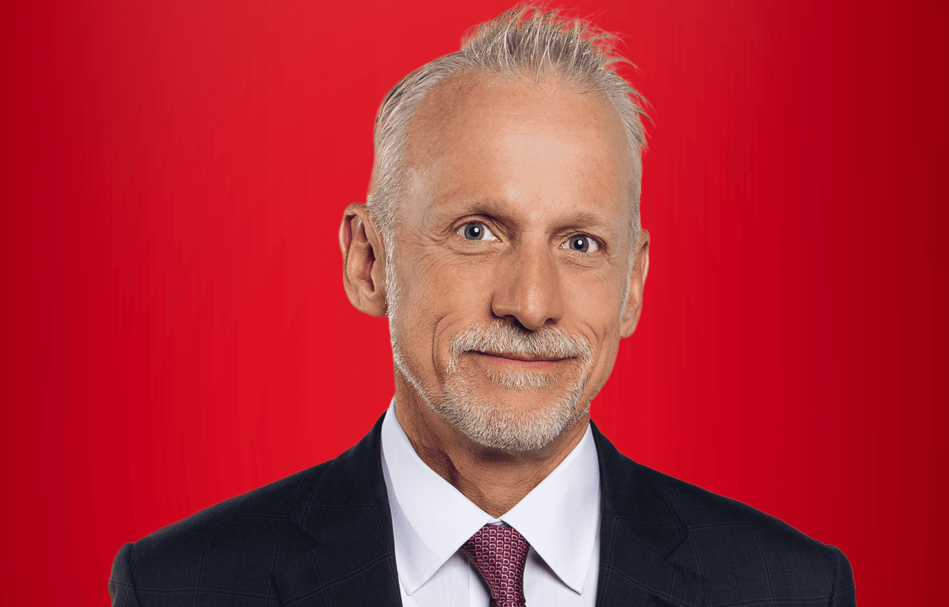 Headshot portrait of Tom McMahon wearing professional attire against a solid red background. 