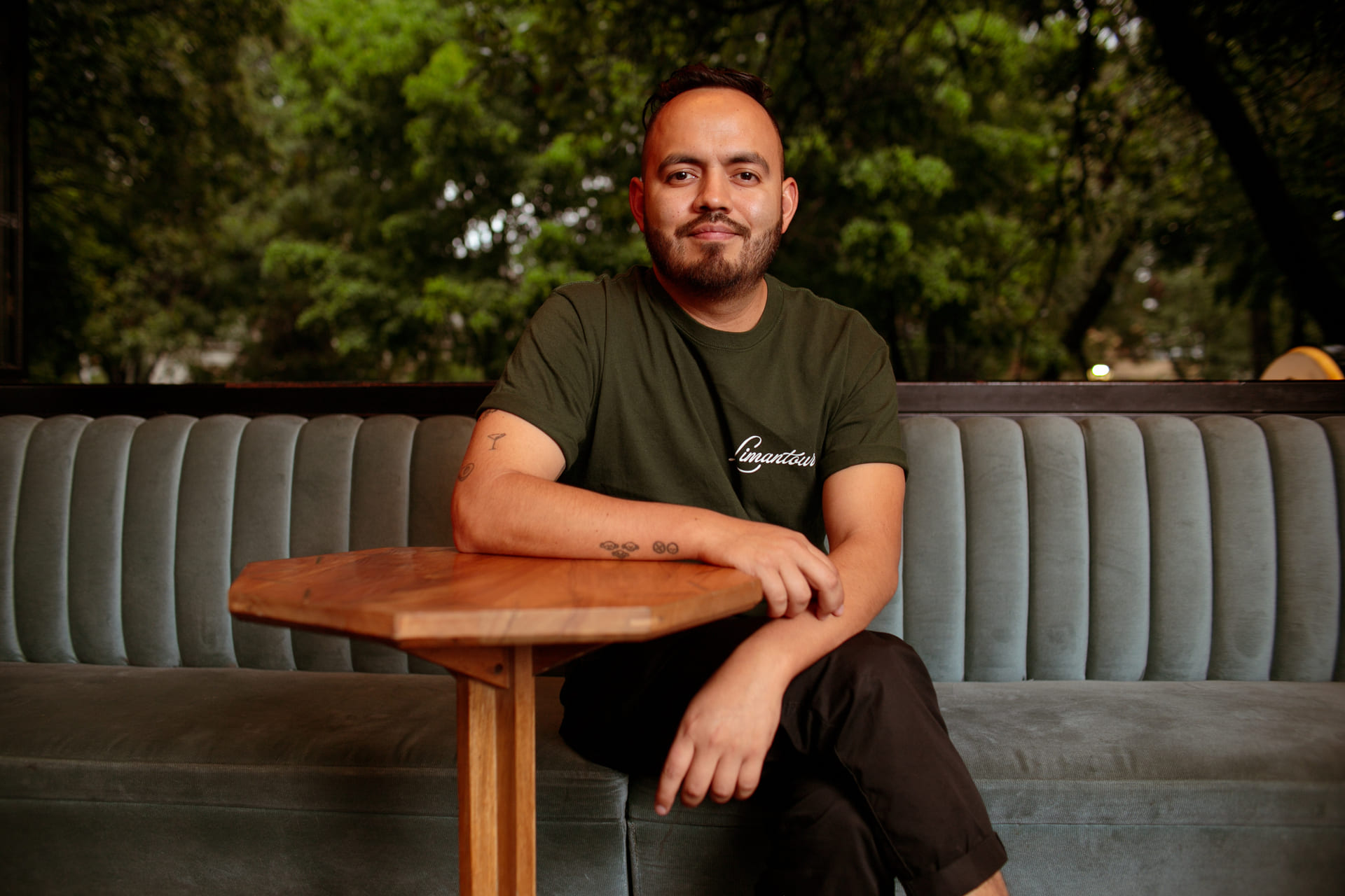 José Luis León Martínez seated at a wooden table in a booth with a green shirt and tattoos, looking at the camera.
