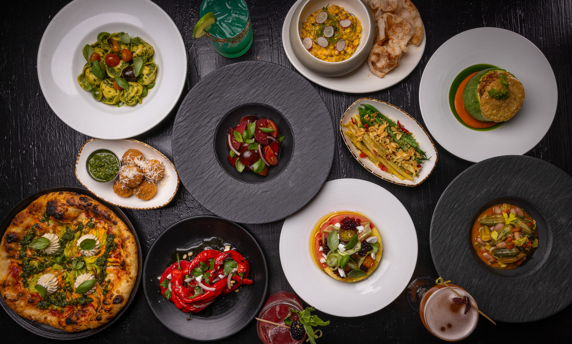 Overhead view of a colorful summer menu featuring gourmet dishes like pasta, pizza, salads, vegetables, and cocktails on a black table.