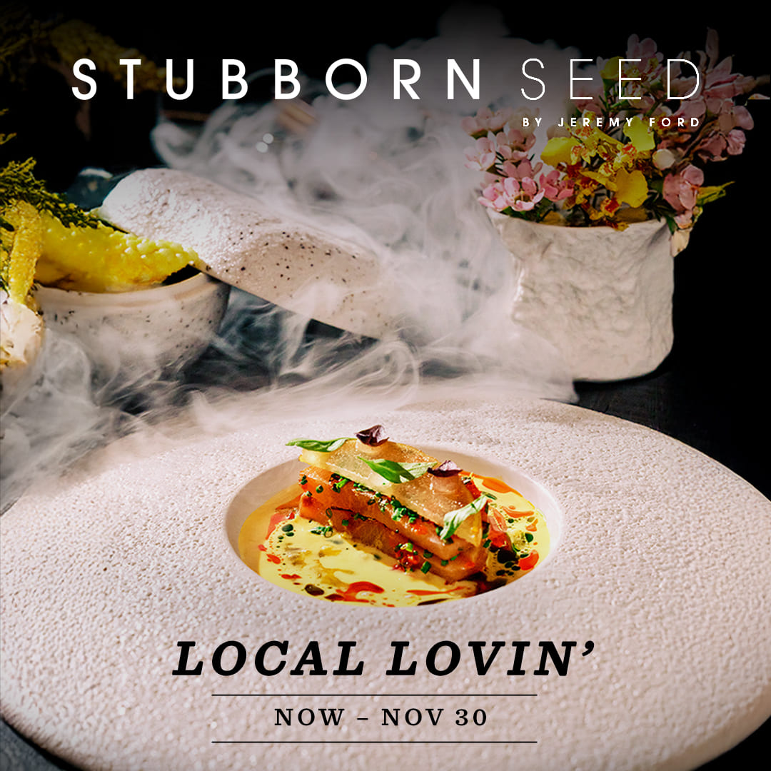 A gourmet dish plated with artistic precision, featuring layers of fresh ingredients in a shallow bowl set within a textured white plate, surrounded by dramatic wisps of smoke. Decorative floral arrangements and crispy garnish accent the background, enhancing the fine dining presentation.