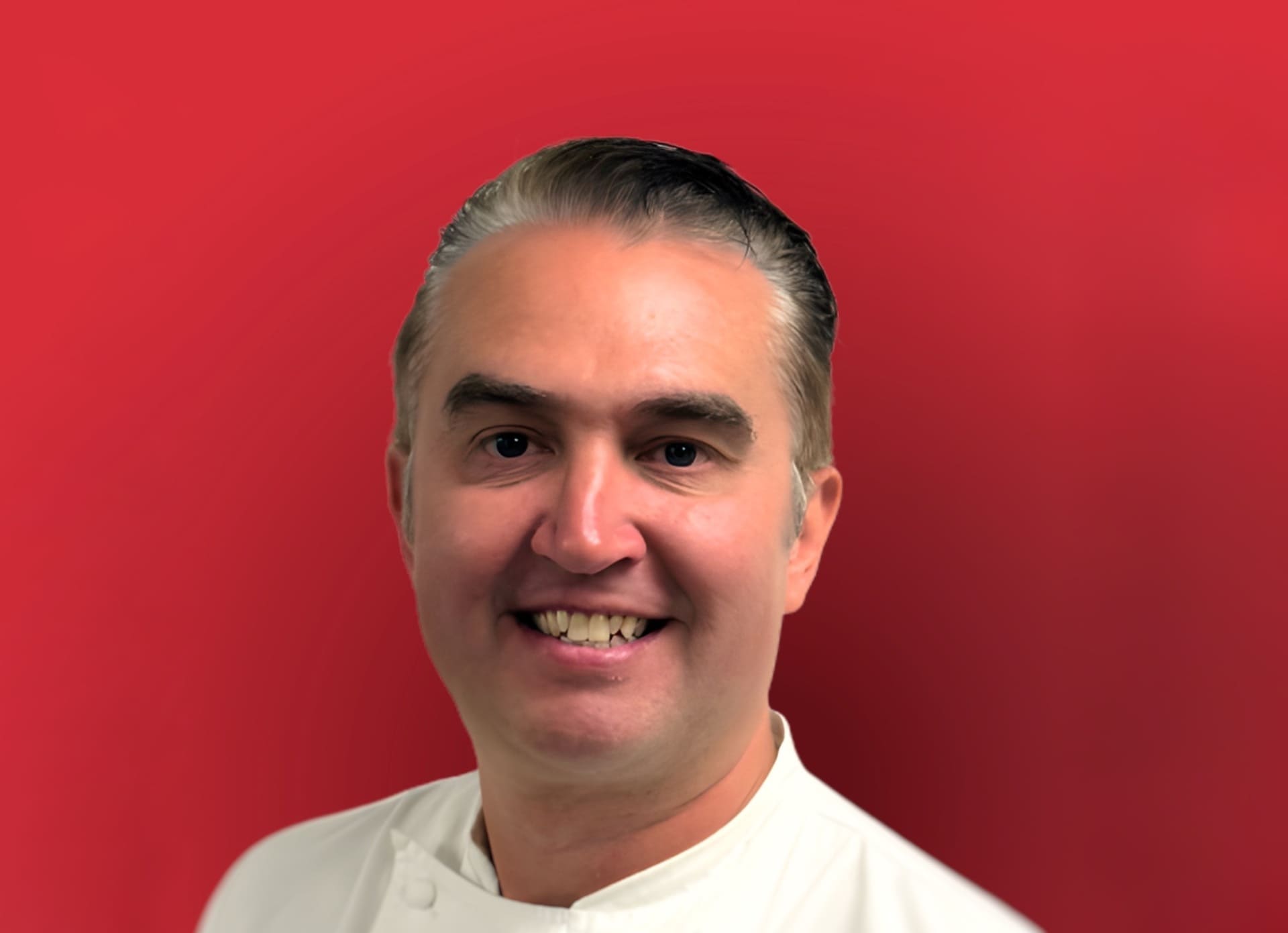 Smiling male chef Lee Bennett in a white uniform standing against a vibrant red background.