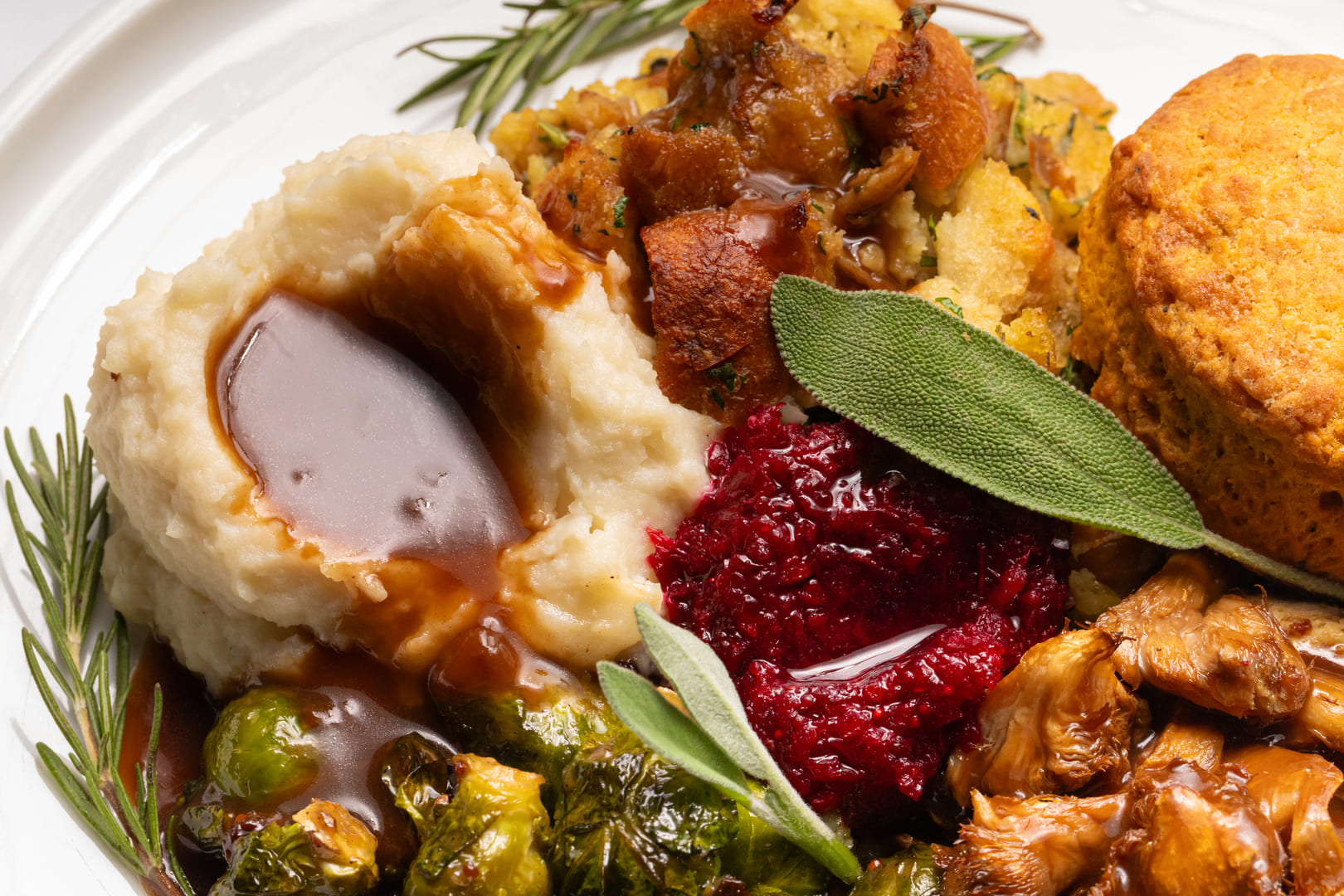 A Thanksgiving-inspired vegan meal featuring creamy mashed potatoes with brown gravy, Brussels sprouts, cornbread stuffing, cranberry relish, sautéed mushrooms, and a golden biscuit, garnished with fresh rosemary and sage.
