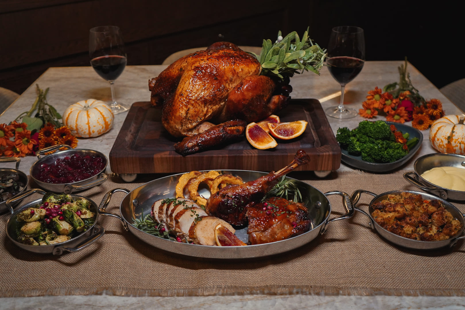 A festive Thanksgiving dinner spread featuring a golden roasted turkey on a cutting board, surrounded by sliced blood oranges, wine glasses, small decorative pumpkins, and various side dishes including mashed potatoes, stuffing, Brussels sprouts, and cranberry sauce, all artfully plated on a fall-themed tablescape.