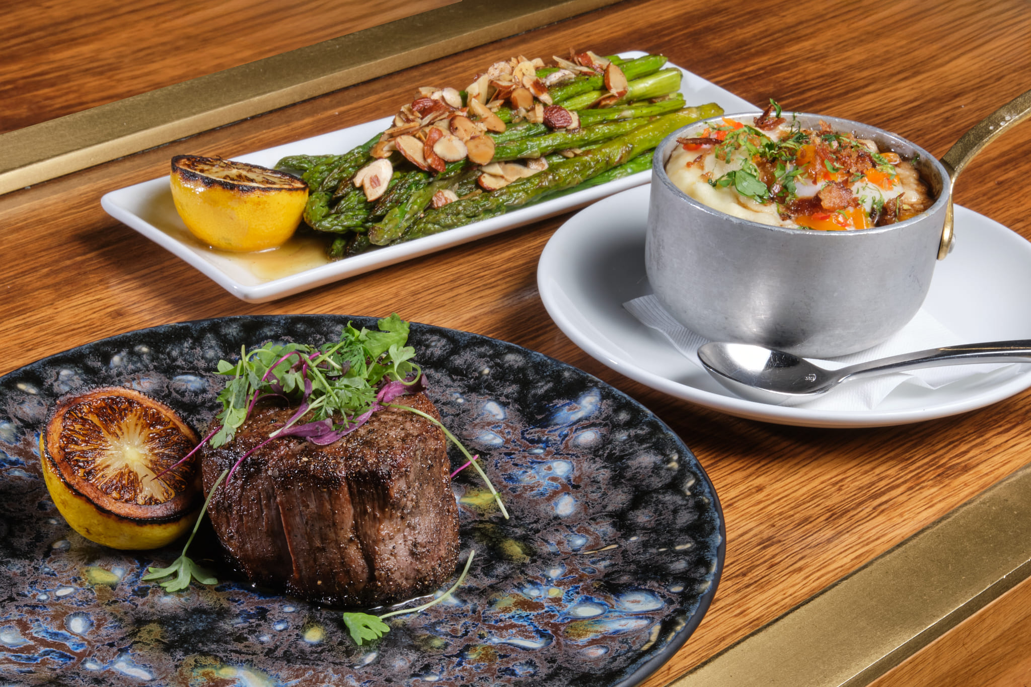 Elegant steak dinner featuring a seared filet mignon with charred lemon, creamy mashed potatoes garnished with herbs and sauce, and roasted asparagus topped with slivered almonds, served on a wooden table.