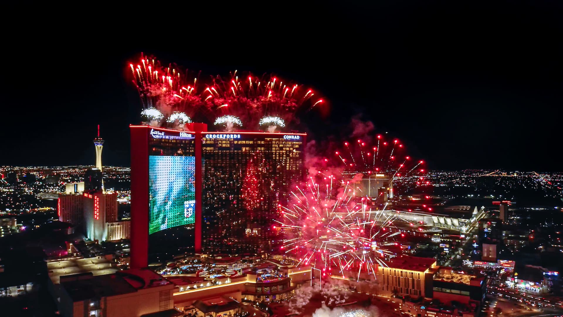 A nighttime aerial view of Resorts World Las Vegas with bright red fireworks exploding above and around the property, lighting up the surrounding Strip.