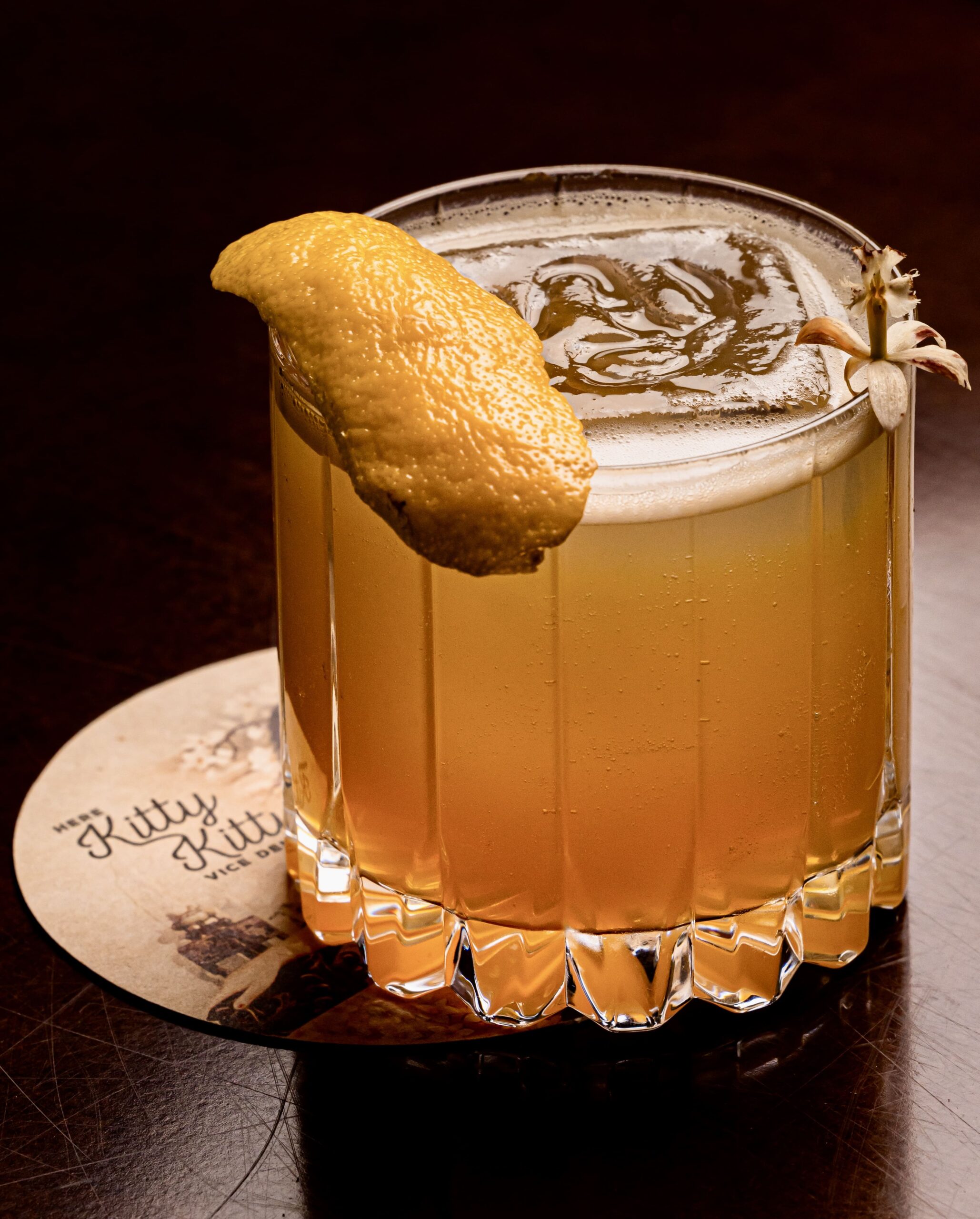 Short glass filled with an amber-colored cocktail, garnished with a lemon peel, resting on a coaster on a dark table.