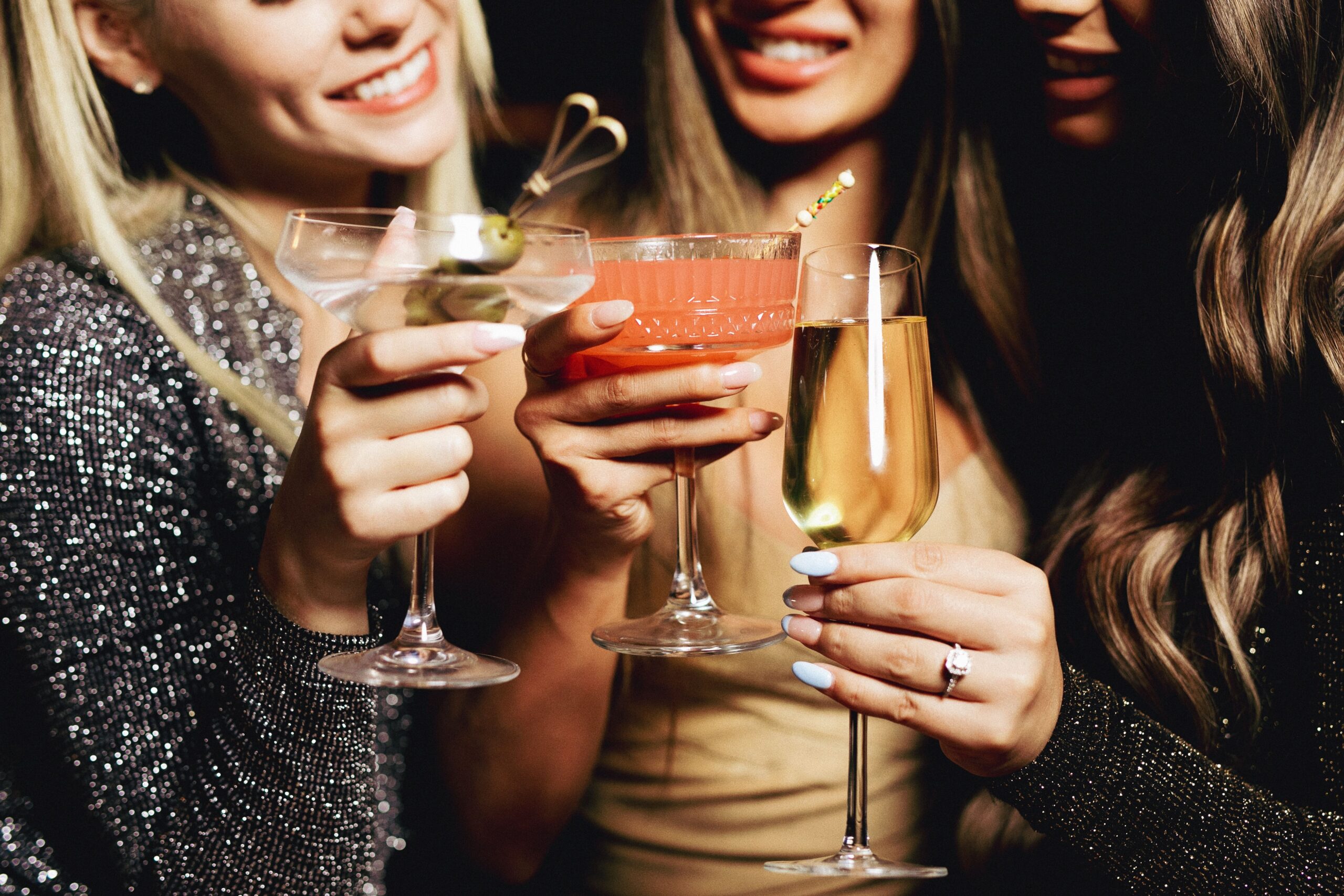 Close-up of three women smiling and raising assorted cocktails and champagne glasses during a celebration, with sparkling outfits and manicured hands visible.