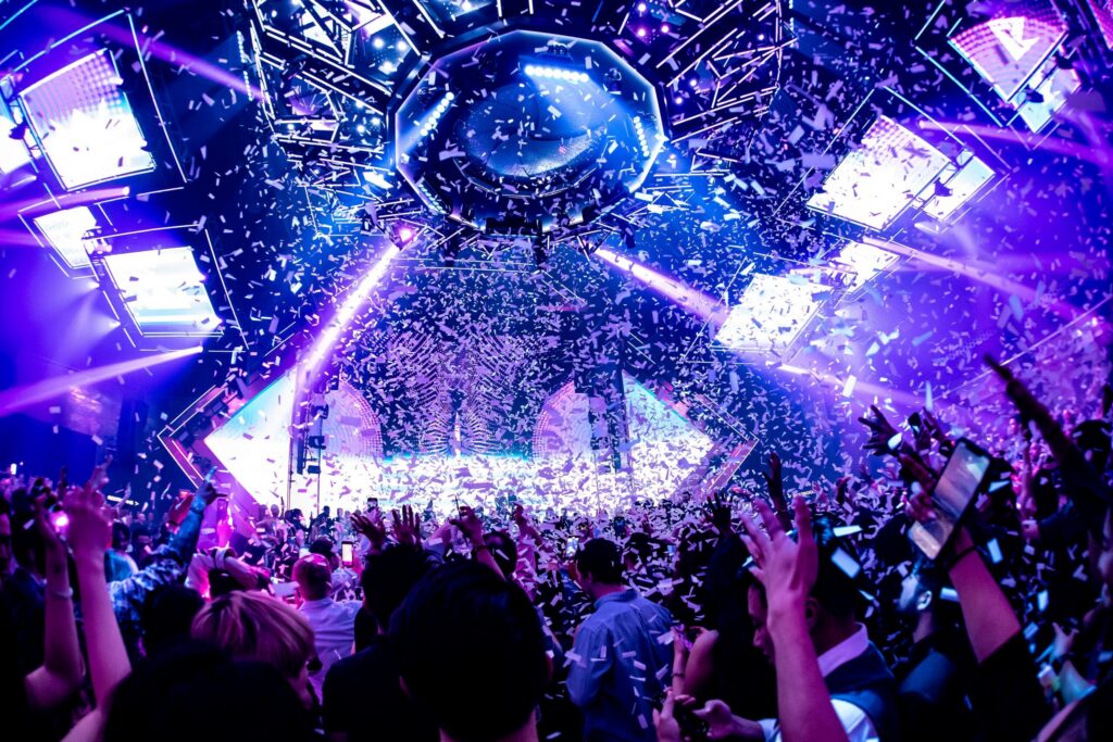 Crowd celebrating on a packed nightclub dance floor as confetti falls under colorful stage lights and LED screens