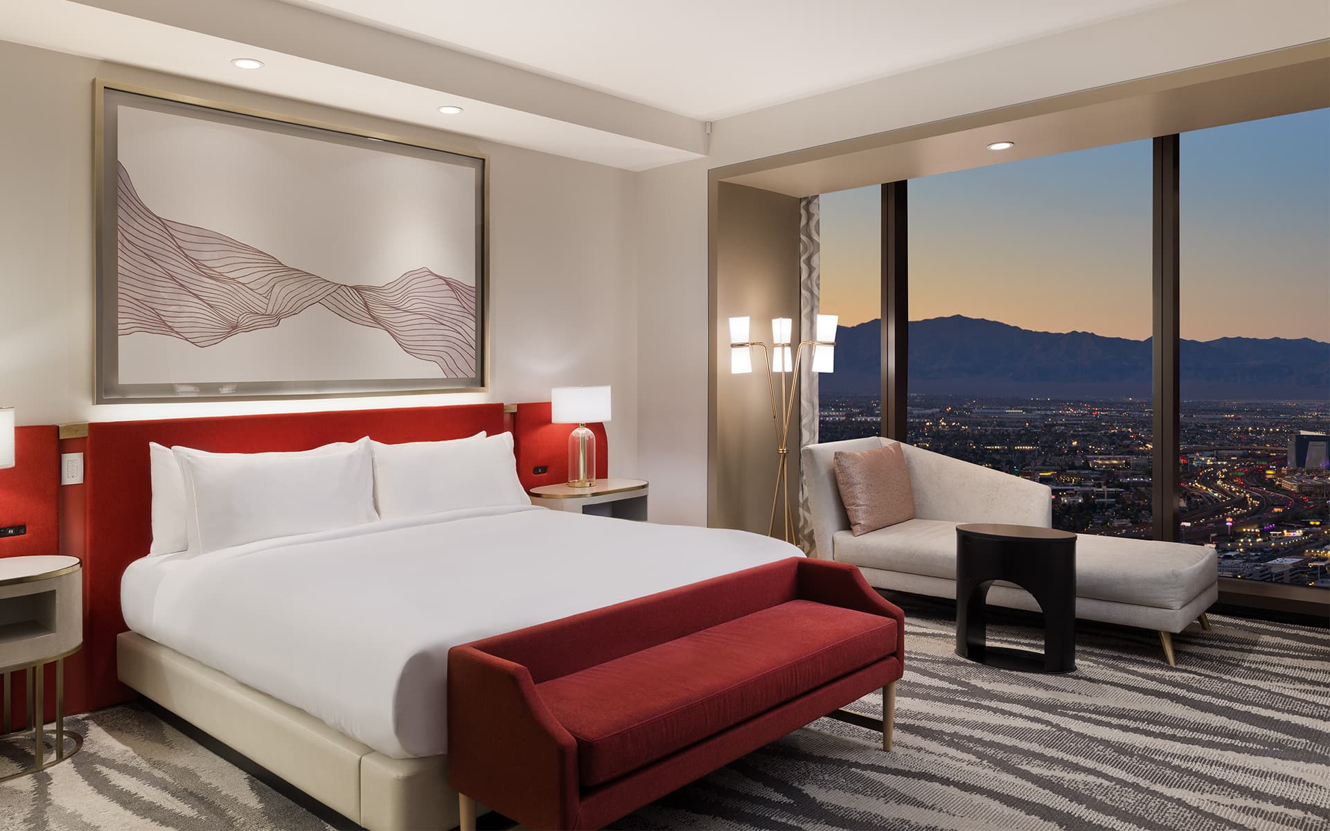 Modern luxury hotel suite with a king bed, red bench seating, lounge chair, and floor-to-ceiling windows overlooking mountains at sunset.
