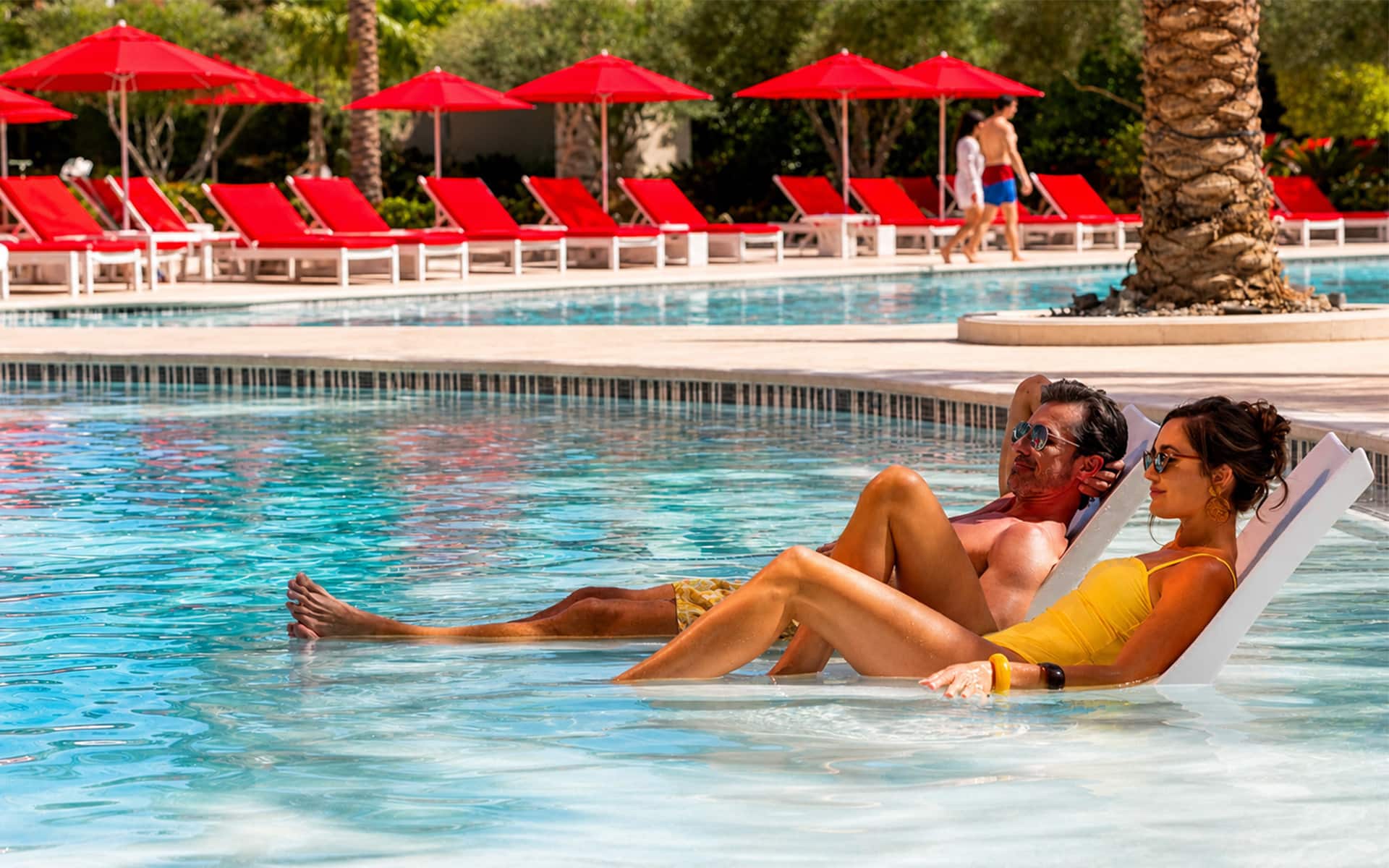 Couple relaxing on in-water lounge chairs at a resort pool surrounded by red umbrellas and palm trees.