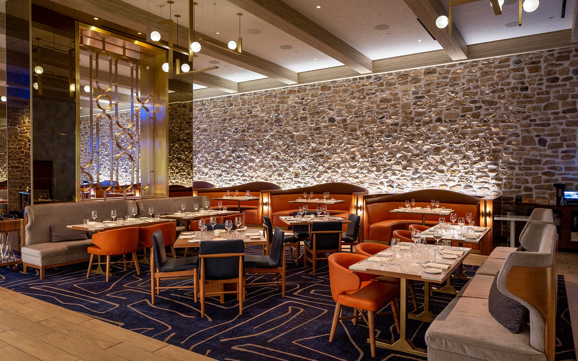 Elegant restaurant dining room with stone accent walls, warm lighting, upholstered seating, and tables set for service.