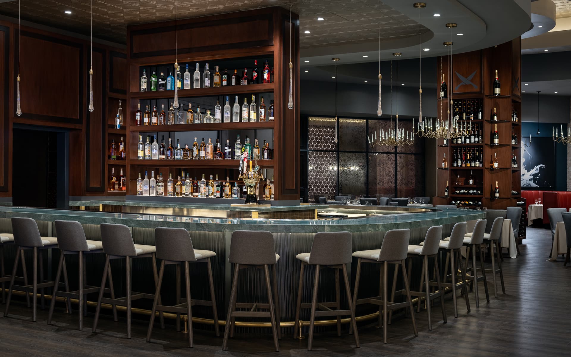 Modern restaurant bar with wraparound seating, backlit liquor displays, wood-paneled finishes, and elegant pendant lighting.