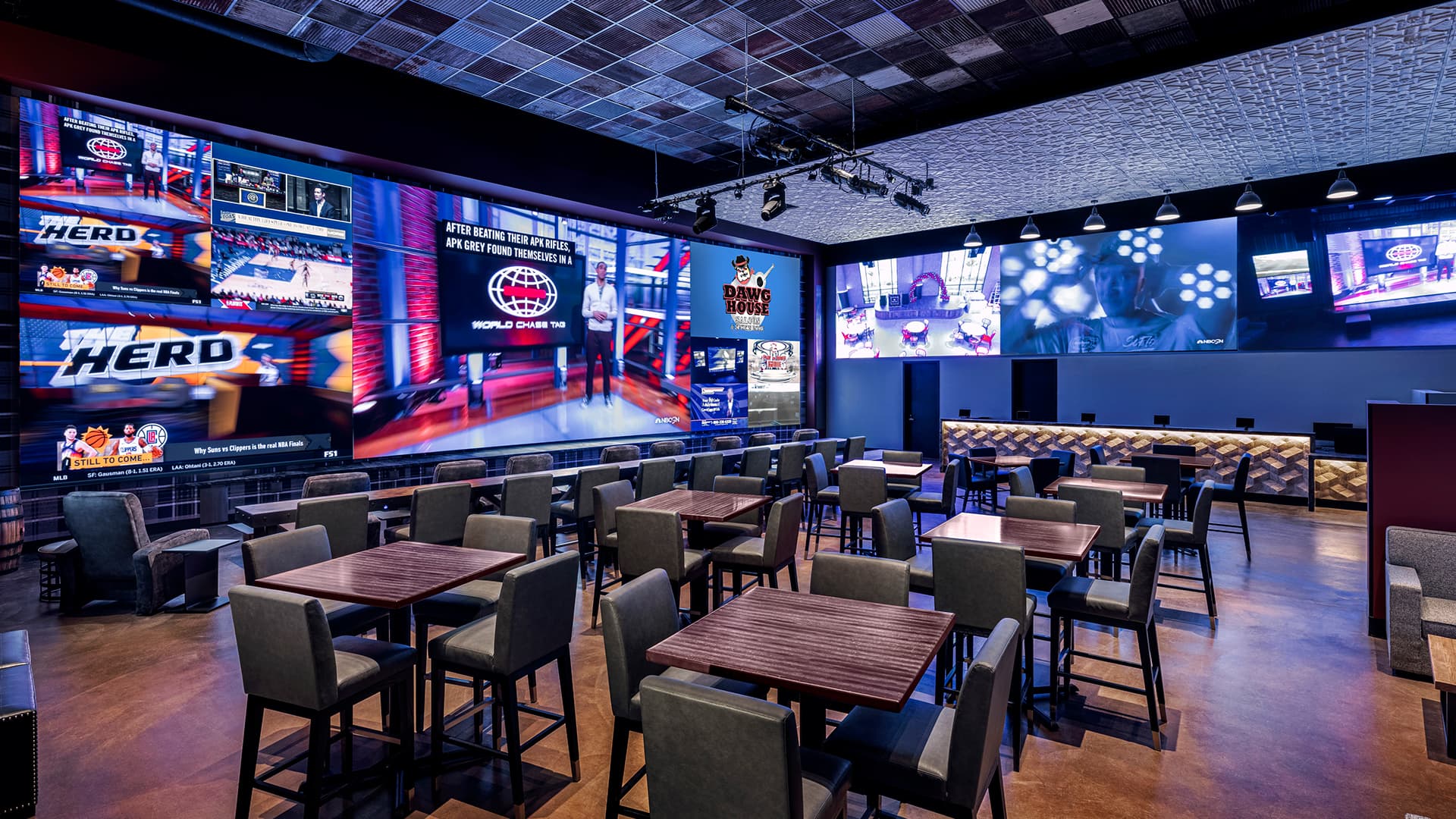 Interior of a sports viewing venue with multiple large video walls displaying live sports content, surrounded by dining tables and seating for guests.