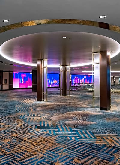 Modern hotel event pre-function space featuring blue patterned carpet, illuminated ceiling details, wood-paneled columns, and digital display screens in the background.