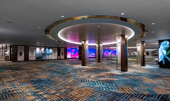 Wide view of a luxury hotel pre-function event space with blue patterned carpet, circular illuminated ceiling feature, wood-paneled columns, and digital display screens.