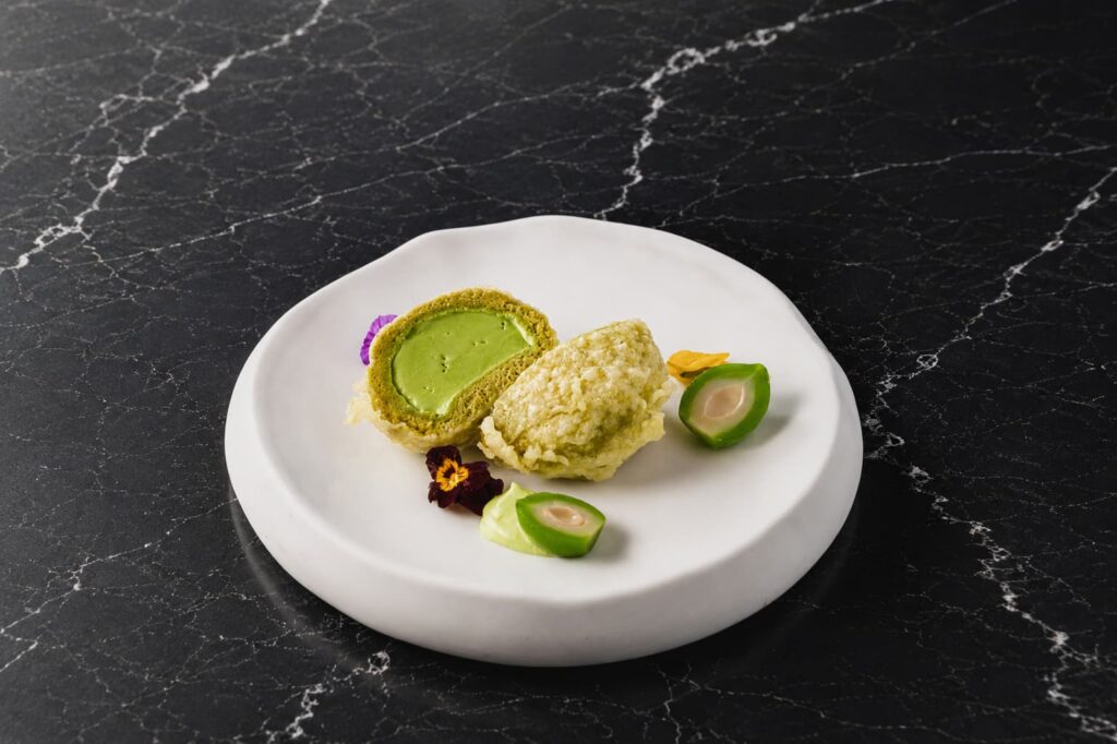 Plated matcha dessert featuring matcha castella cake, wagyu tallow crisp, matcha cream, gelato, and garnishes on a white plate against a black marble surface.