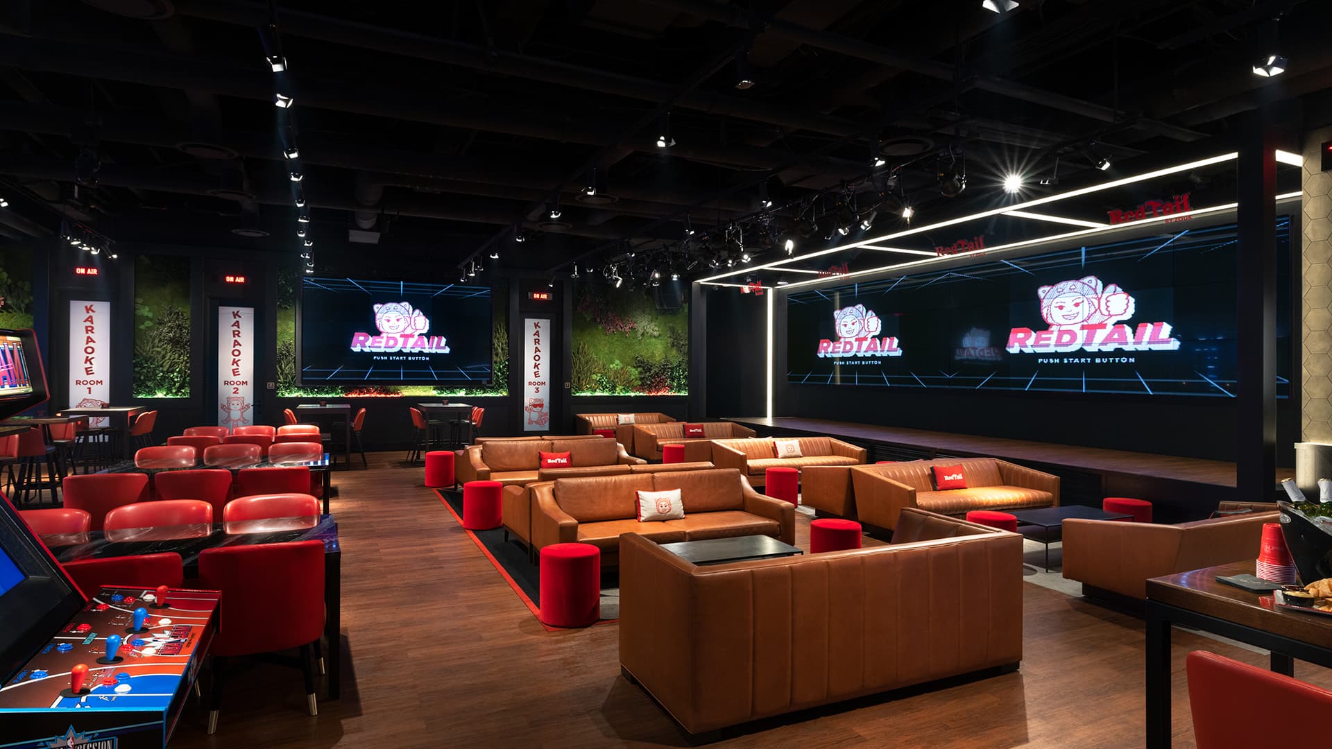 Interior of RedTail featuring lounge seating, gaming tables, arcade games, karaoke rooms, and a large stage with digital screens in a modern social entertainment venue.