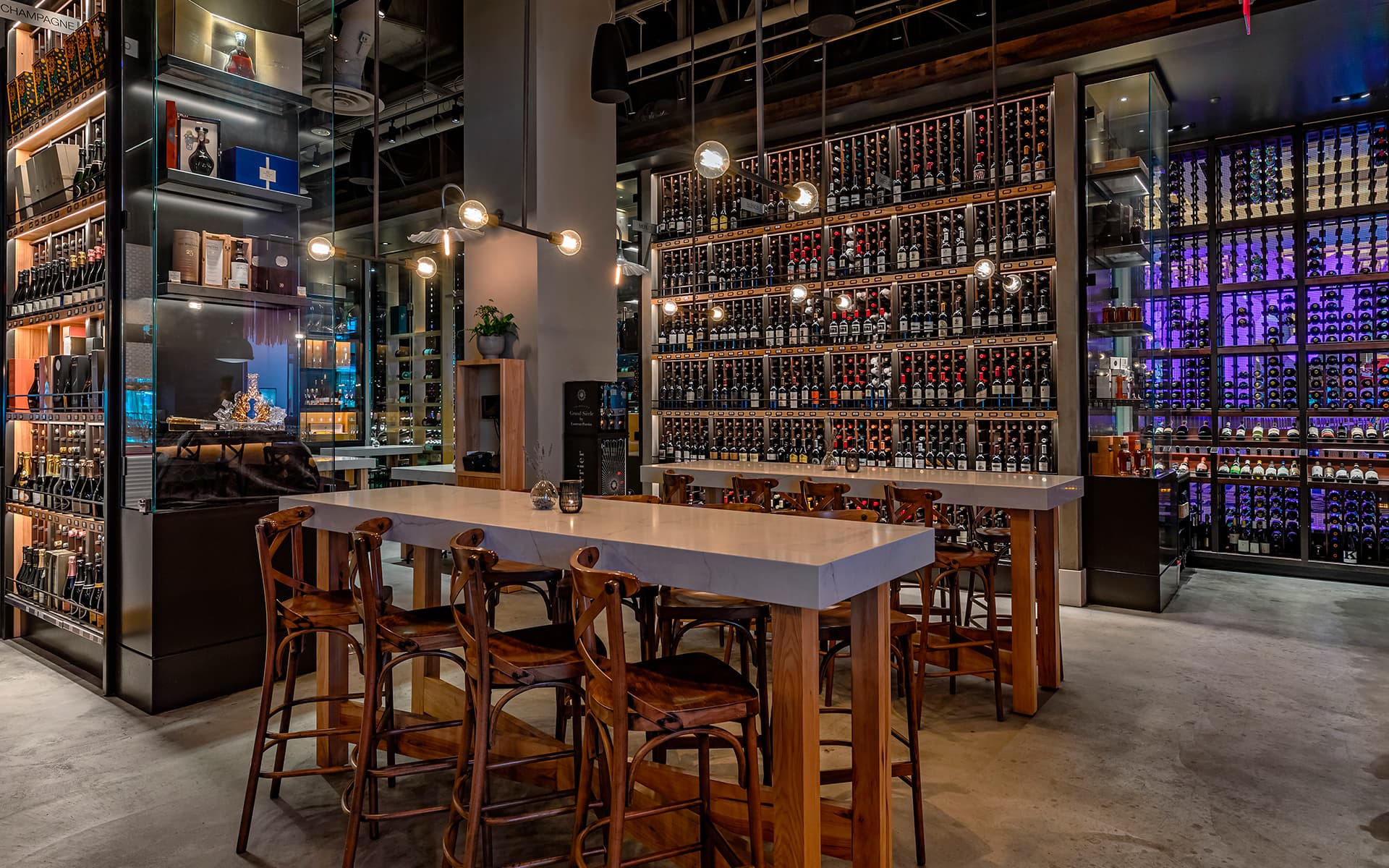 Elegant wine bar interior featuring floor-to-ceiling wine displays, communal dining tables, warm pendant lighting, and contemporary upscale décor.