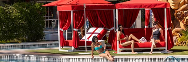 Guests relax in red poolside cabanas beside a resort swimming pool, with one guest sitting at the pool's edge and others lounging inside the shaded cabanas.