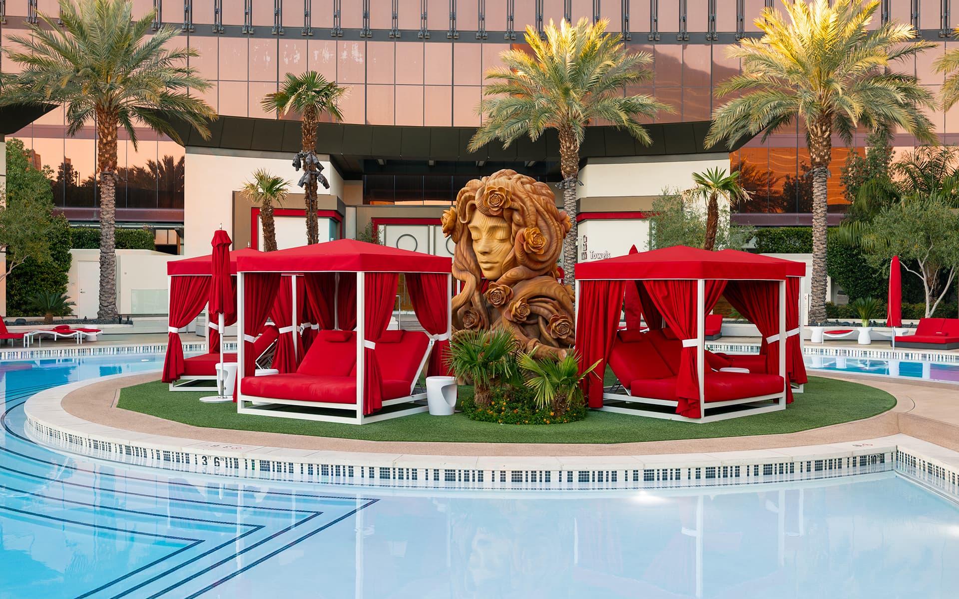 Red pool cabanas surrounding a floral-inspired sculpture beside the pool at Resorts World Las Vegas, with palm trees and the hotel tower in the background.