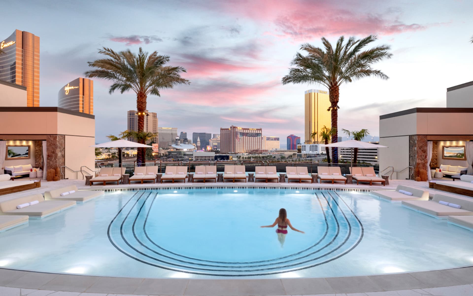 A guest relaxes in the Athena rooftop pool at sunset, with palm trees, lounge chairs, private cabanas, and panoramic views of the Las Vegas Strip under a pink and blue evening sky.