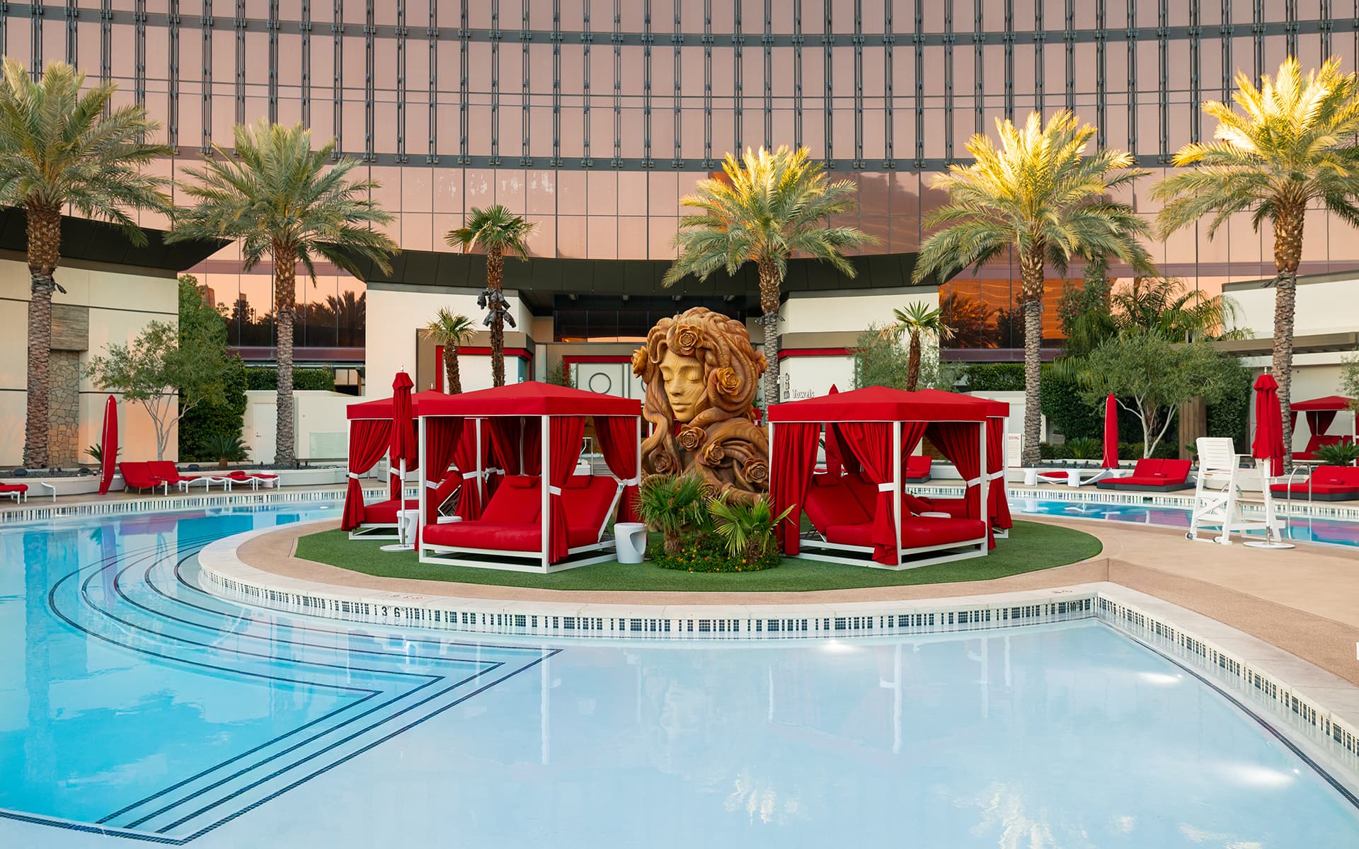 Resort pool surrounded by palm trees, red daybeds and cabanas, with a decorative sculpture at the center.