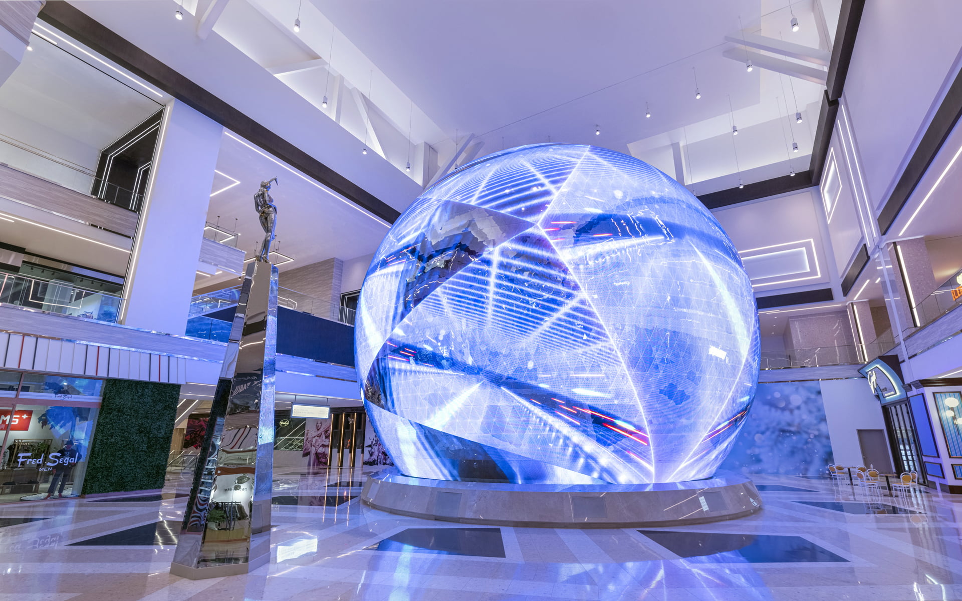 A wide shot of a multi-story indoor shopping mall atrium featuring a large, glowing blue LED sphere sculpture with geometric patterns as the central installation.
