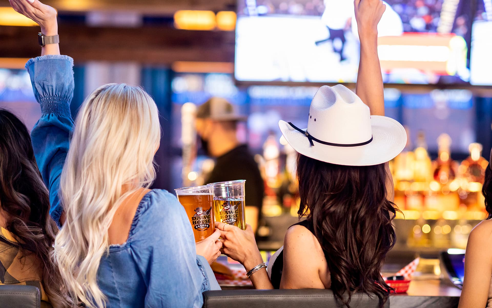 Guests raise their hands and toast with beers while watching screens at a lively bar.