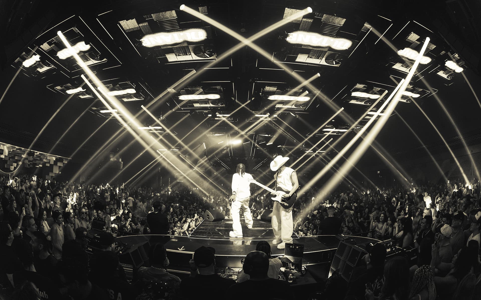 Two performers on stage surrounded by a large crowd as dramatic beams of light illuminate the venue.