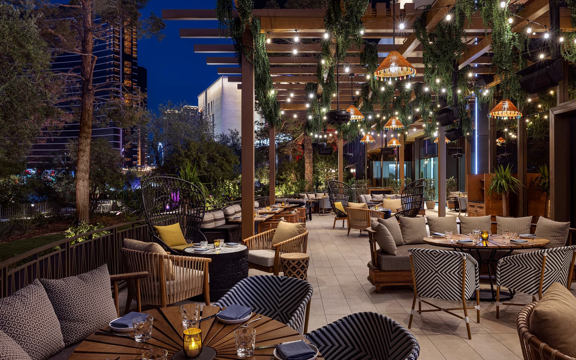 Outdoor dining patio with set tables, lounge seating, hanging greenery, and string lights at night.
