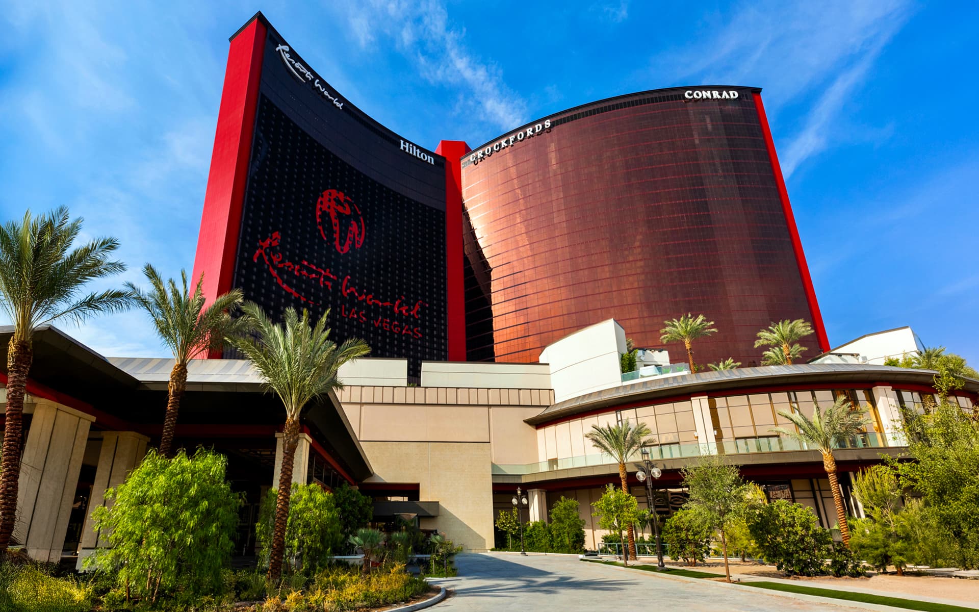Exterior view of Resorts World Las Vegas with its red hotel towers and palm trees under a blue sky.
