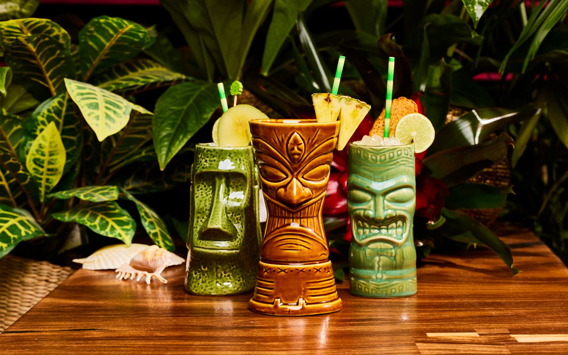 Three tropical cocktails served in tiki-style ceramic mugs with fruit garnishes on a wooden table surrounded by lush green plants.