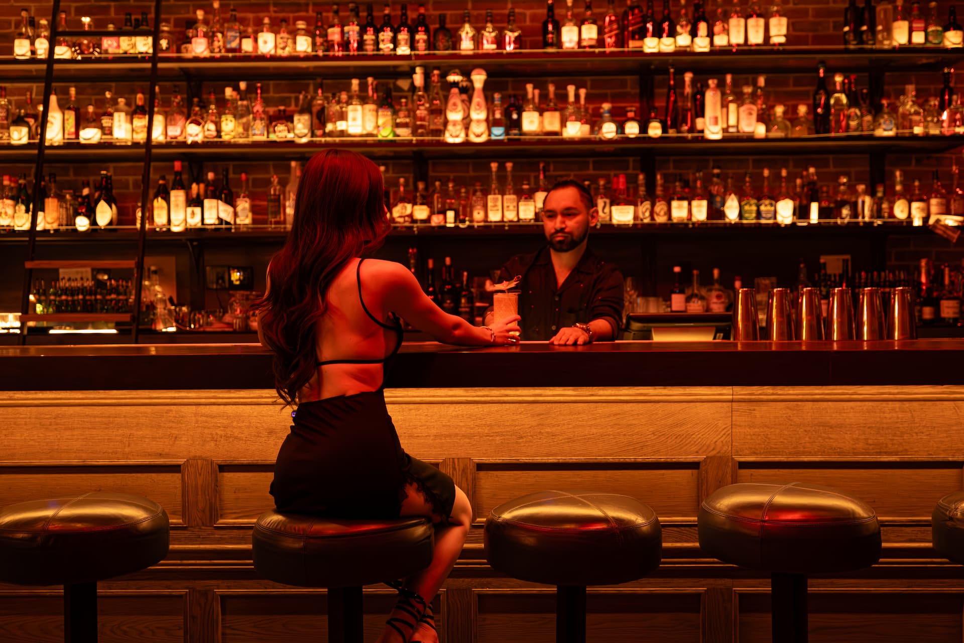 Woman seated at a warmly lit bar receiving a cocktail from a bartender.