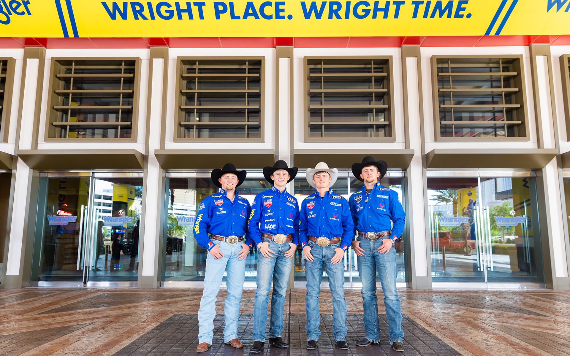 Four rodeo cowboys in matching blue shirts and cowboy hats pose together outside a Wrangler-branded entrance.