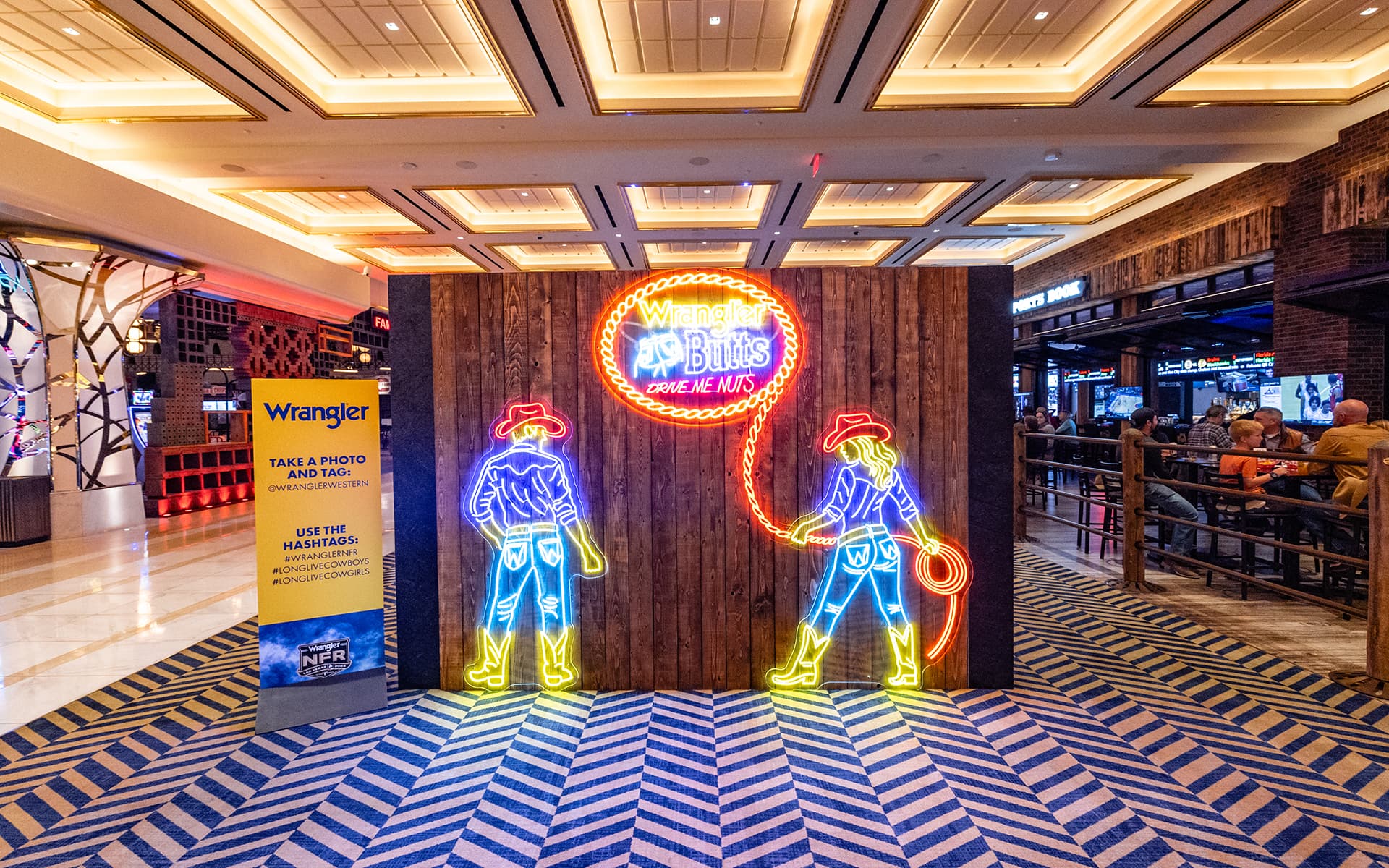Colorful neon cowboy and cowgirl figures illuminate a Wrangler rodeo-themed photo display with a rustic wood backdrop.