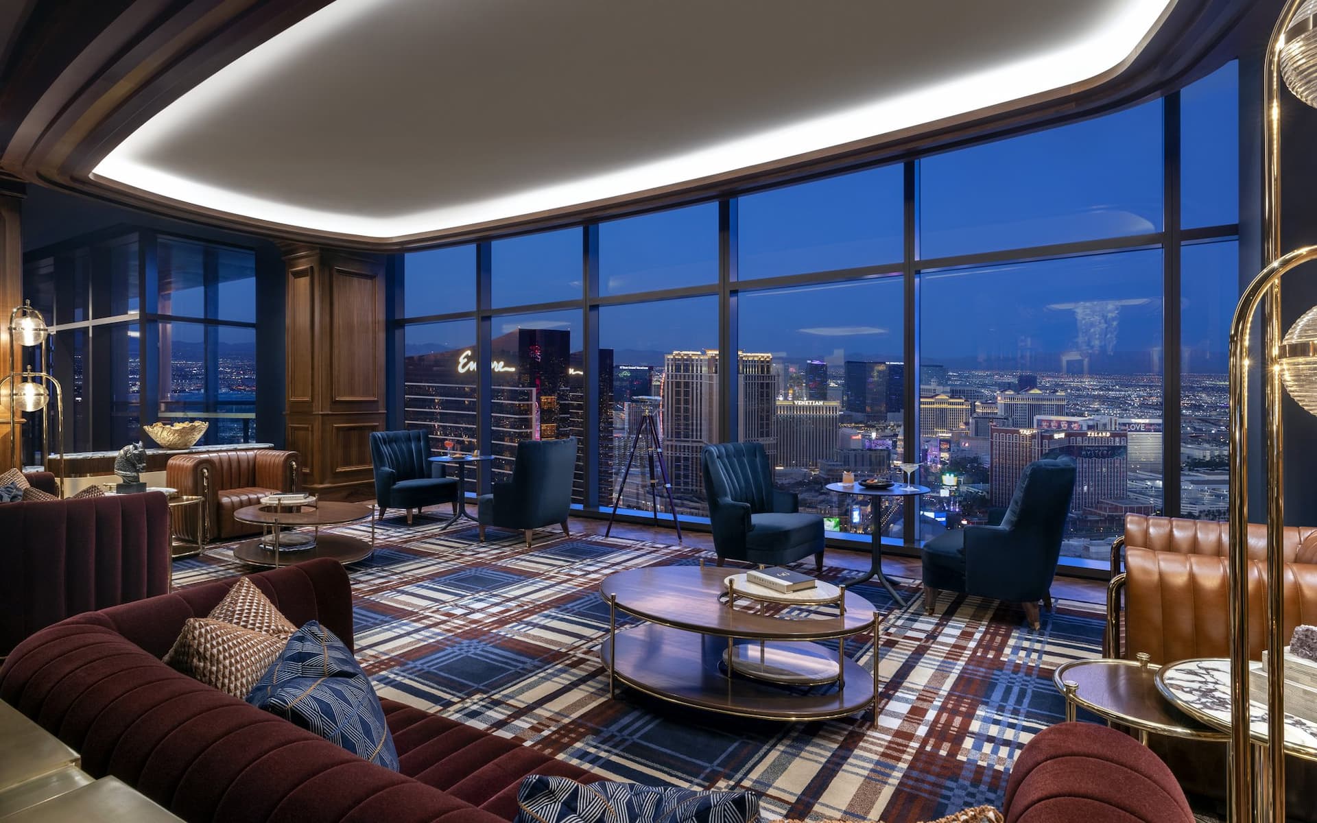 Elegant interior of Allē Lounge on 66 featuring plush sofas, blue armchairs, plaid carpeting, and floor-to-ceiling windows overlooking the Las Vegas Strip at dusk.