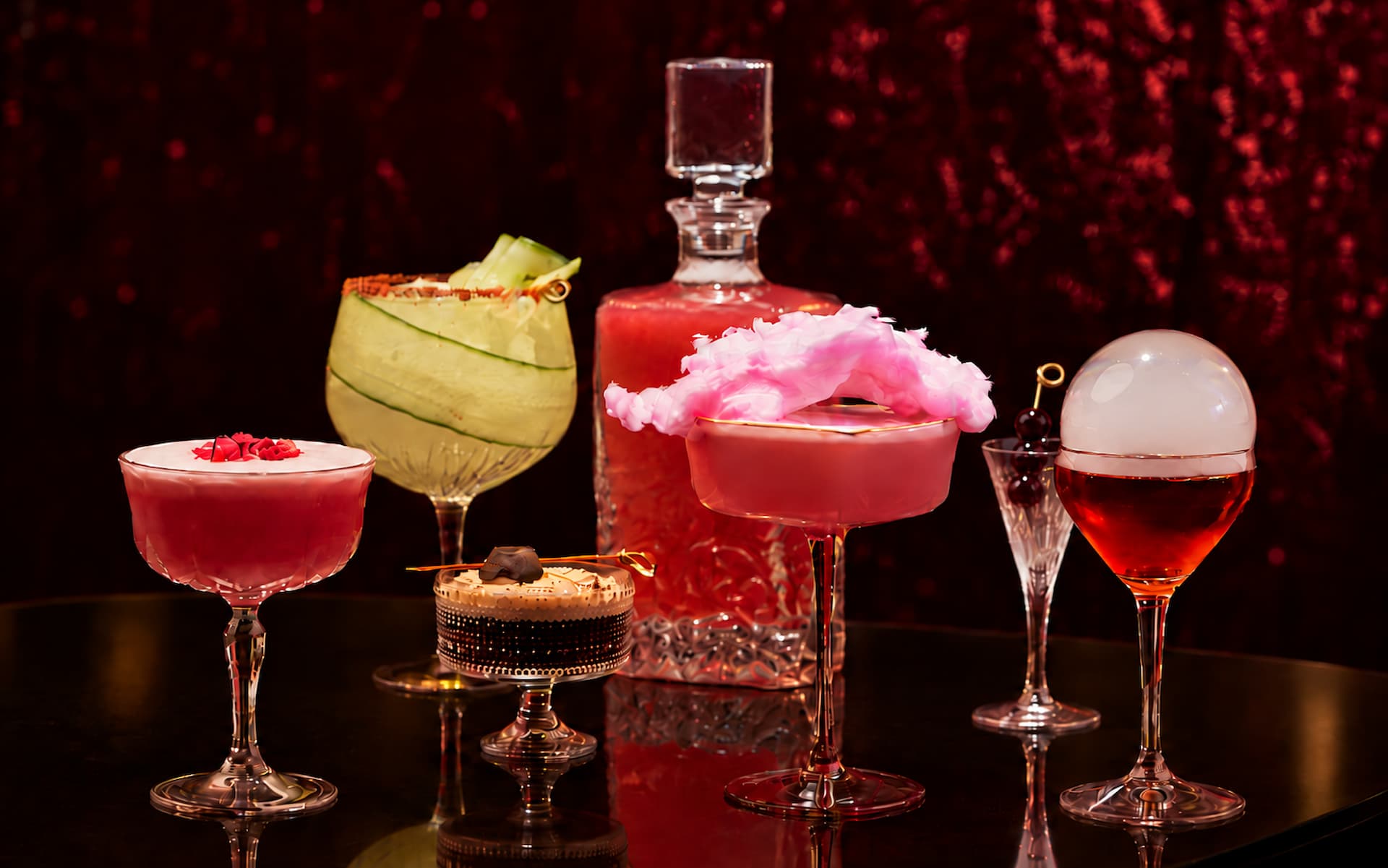 An elegant collection of colorful signature cocktails in decorative glassware, featuring pink, red, and green drinks with creative garnishes, arranged against a rich burgundy velvet backdrop.