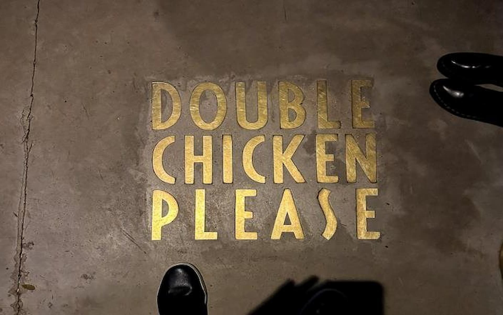 Gold lettering spelling "Double Chicken Please" on a concrete floor, with black shoes partially visible around the edges.