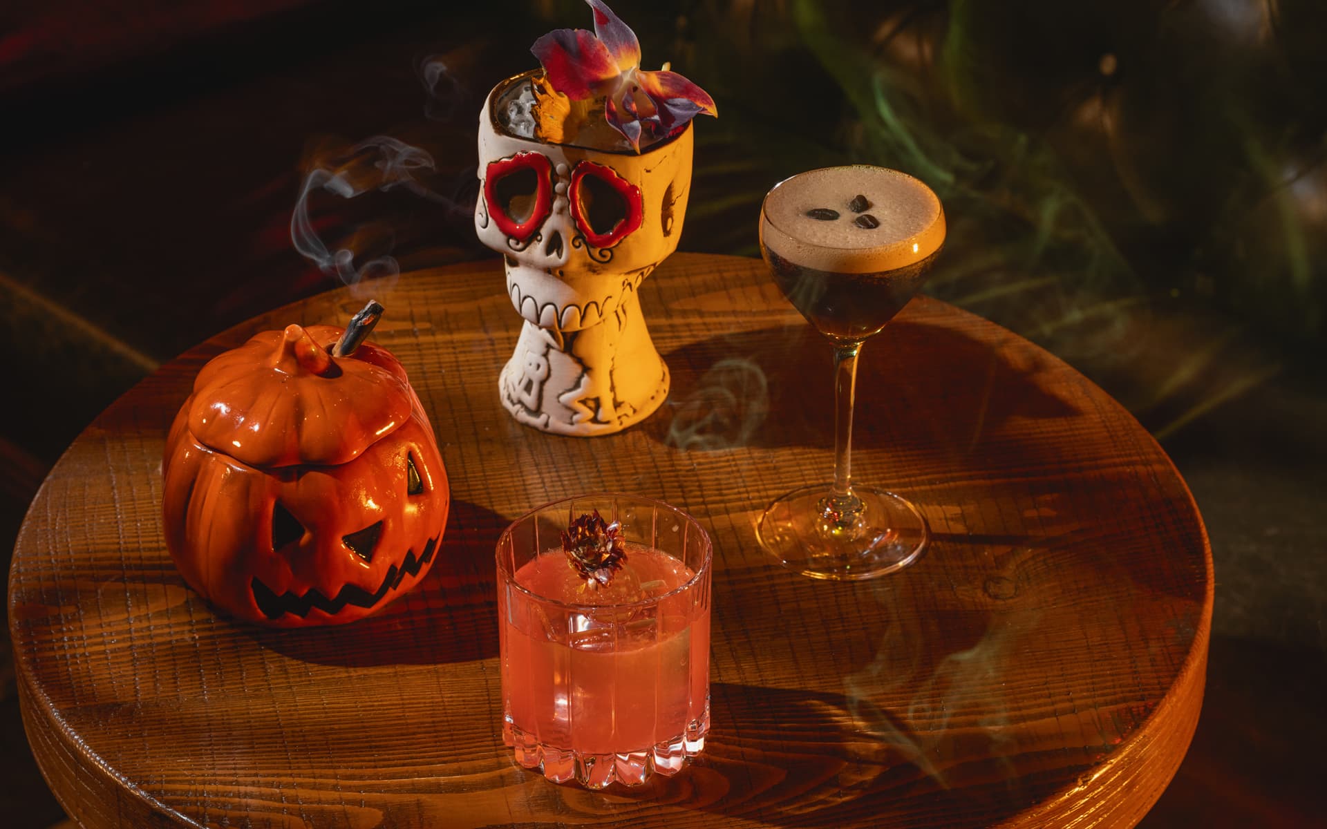 Three Halloween-themed cocktails displayed on a wooden table with a jack-o’-lantern and decorative skull amid wisps of smoke.