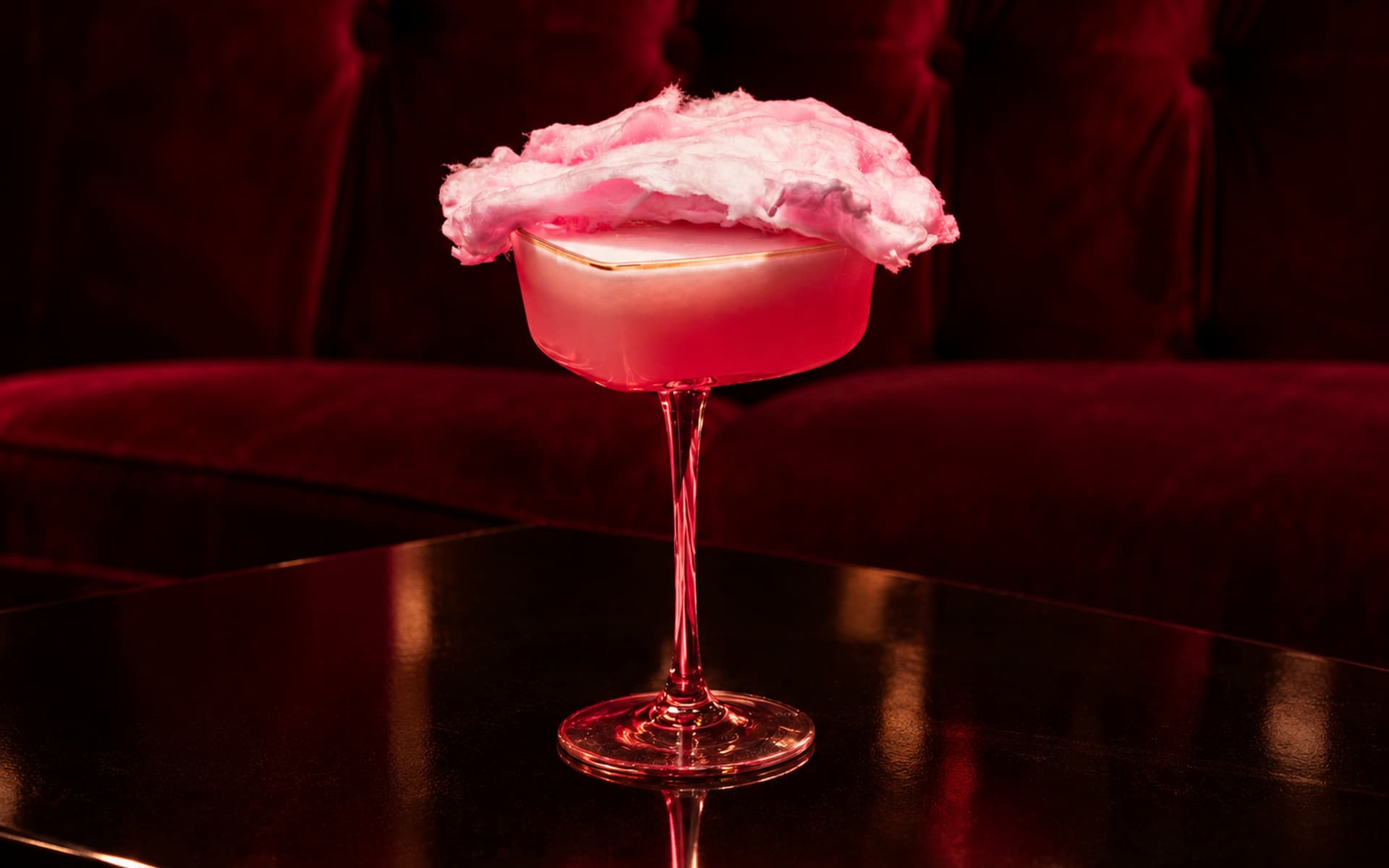 Pink cocktail served in an elegant stemmed coupe glass, topped with fluffy pink cotton candy, displayed on a dark reflective table against a deep burgundy velvet backdrop.