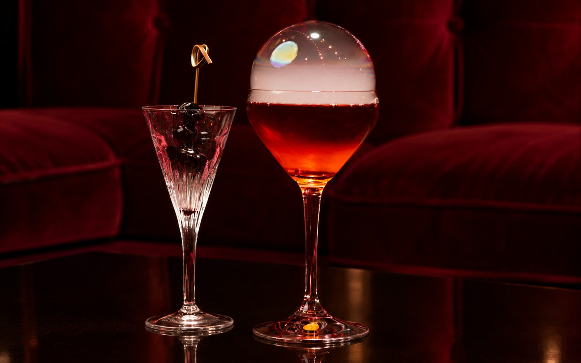 Red signature cocktail served in an elegant stemmed glass topped with a translucent smoke bubble, accompanied by a crystal glass filled with dark cherries, against a burgundy velvet backdrop.